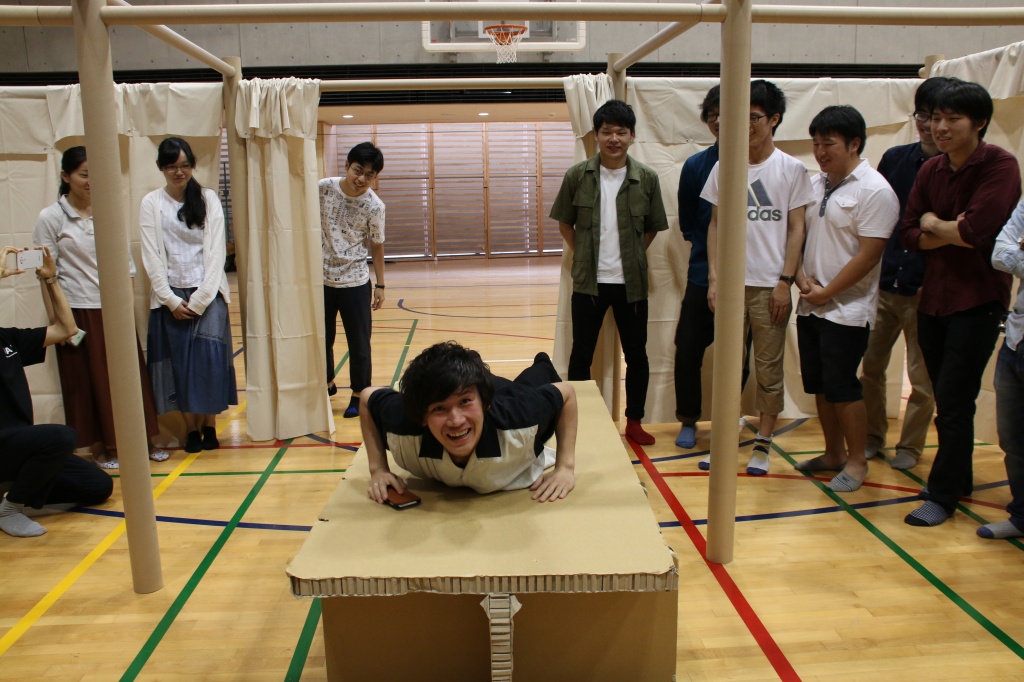 仮設間仕切りの中に組まれた段ボールベッドに乗って感触を確かめる学生